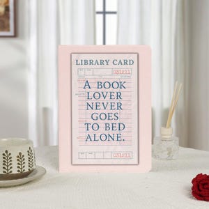 Puede incluir: Una impresión enmarcada en rosa con un diseño de tarjeta de biblioteca vintage y la cita "A Book Lover Never Goes to Bed Alone" en texto azul. La impresión está sobre una superficie blanca con una taza y un difusor decorativos.