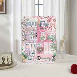 May include: An art print depicting a charming European street scene with pink buildings, flowering trees, and a vintage pink convertible car. The artwork has a soft, pastel colour palette.