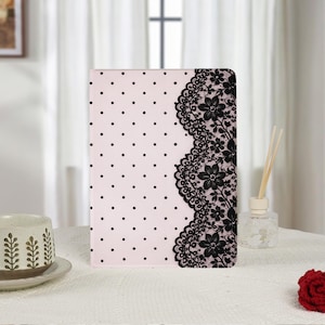 Puede incluir: Una funda rectangular para tableta con fondo rosa claro, un patrón de lunares negros en el lado izquierdo y una franja vertical ancha de intrincado encaje floral negro a lo largo del borde derecho de la cubierta frontal.