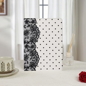 Puede incluir: Una funda rectangular para tableta con fondo blanco, un patrón de puntos negros en el lado derecho y una franja vertical de intrincado encaje floral negro a lo largo del borde izquierdo, cubriendo el área del lomo de la cubierta protectora.