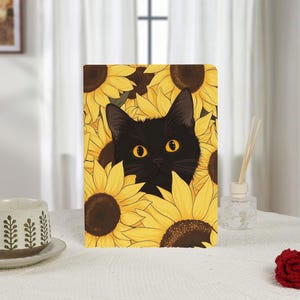 May include: A rectangular notebook featuring a black cat peeking out from a field of yellow sunflowers. The cat has bright yellow eyes and black fur. The sunflowers have brown centers and yellow petals. The notebook is on a white surface.