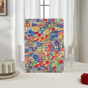 May include: A floral patterned journal with a vibrant mix of red, blue, yellow, and orange flowers on a cream background. The notebook is rectangular and stands upright on a white surface. The design is reminiscent of a botanical garden.