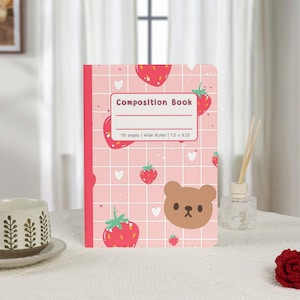 May include: A pink composition book with a strawberry and bear pattern. The cover features the text "Composition Book" and "110 pages | Wide Ruled | 7.5 x 9.25". The notebook is on a white surface.