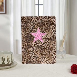 Può includere: Un quaderno con una copertina con stampa leopardata e una stella rosa al centro. La stella presenta anche un motivo leopardato. Il quaderno è appoggiato su una superficie bianca.