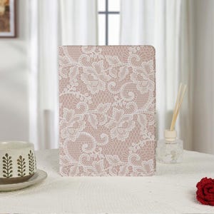 Puede incluir: Una funda rectangular para tableta o libro con un fondo rosa empolvado cubierto por un detallado patrón de encaje floral blanco. La funda se mantiene en posición vertical, mostrando un diseño continuo de flores y malla entrelazada.
