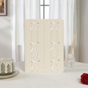 May include: Cream-coloured notebook featuring a cable knit pattern and six decorative bows. The cover has a textured appearance, with the bows adding a delicate touch. The notebook is placed on a white surface.