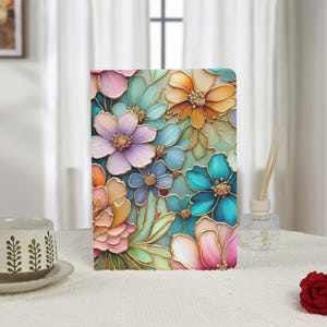 May include: A floral notebook featuring a vibrant design of colorful flowers in shades of pink, orange, blue, and purple with gold outlines. The notebook is rectangular and stands upright on a white surface.