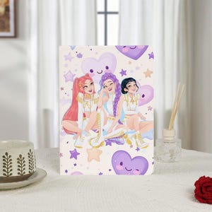Può includere: Un taccuino decorativo con un'illustrazione di tre donne sorridenti con i capelli lunghi, vestite con abiti bianchi e dorati. Lo sfondo è chiaro con cuori e stelle viola.