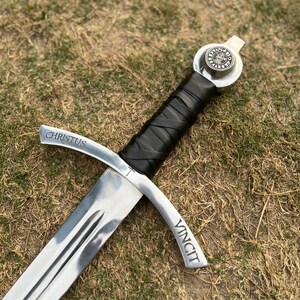 May include: A silver sword with a black leather-wrapped handle, lying on grass. The crossguard is inscribed with "CHRISTUS" and "VINCIT". The pommel features a circular emblem. The blade is polished and reflects light.