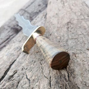 Puede incluir: Un cuchillo artesanal con una hoja ondulada y un mango de madera. El cuchillo presenta una guarda de latón y un mango de madera detallado. La hoja es plateada y el mango es una mezcla de tonos marrones y grises. El cuchillo está diseñado para utilidad y exhibición.