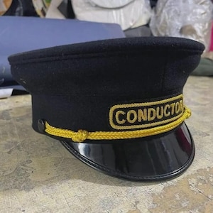 Op de afbeelding: Zwarte conducteurshoed met een glanzende zwarte klep en gouden afwerking. De hoed heeft het woord "CONDUCTOR" geborduurd in gouddraad. Een klassiek accessoire.