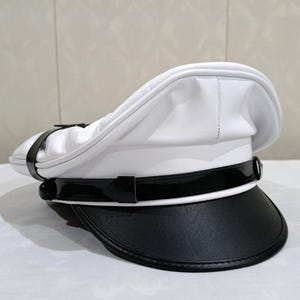 以下が含まれることがあります： 黒いバイザーとバンドが付いた白い船長の帽子。 帽子は丸みを帯びたクラウンと光沢のある黒いブリムを備えています。 滑らかな、おそらく革のような素材で作られています。