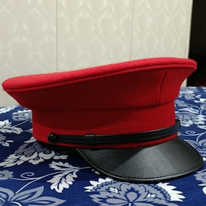 May include: A red wool hat with a black leather visor. The hat features a black band around the base and a button detail. The hat has a classic military or conductor style.