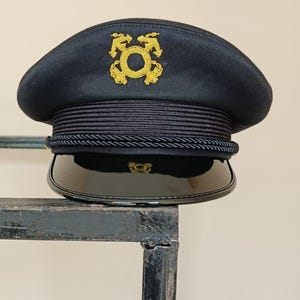 Op de afbeelding: Een marineblauwe hoed met een gouden embleem. De hoed heeft een zwarte rand en een decoratieve touwband. De hoed rust op een metalen oppervlak. De hoed lijkt van militaire of nautische stijl te zijn.