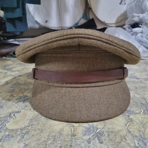 May include: A brown military-style hat with a rounded crown and a short, curved brim. The hat features a dark brown leather band around the base of the crown. The hat is made of a textured, woven fabric.