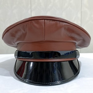 Può includere: Un cappello in pelle marrone con una tesa nera lucida. Il cappello presenta una corona arrotondata e una fascia nera sopra la tesa. Il cappello è probabilmente di stile militare o formale.