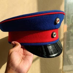 Op de afbeelding: Een blauwe en rode militaire hoed met een zwarte klep. De hoed heeft een blauwe kroon met rode rand en een rode band. Twee zilveren knopen sieren de zijkant. De hoed wordt door een hand vastgehouden.