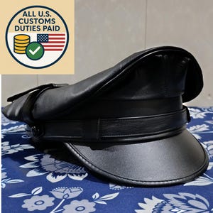 Puede incluir: Un sombrero de cuero negro con una corona redondeada y un ala rígida y curvada. El sombrero presenta un detalle de correa alrededor de la corona y un cierre de hebilla. La imagen también incluye un gráfico circular con el texto "ALL U.S. CUSTOMS DUTIES PAID".