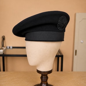 May include: A black beret featuring a structured crown and a wide, flat-brimmed base. The hat is adorned with a circular, pleated fabric rosette detail on the side and includes a thick, textured black grosgrain ribbon band encircling the lower edge.