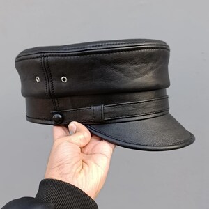 May include: A black leather military-style cap featuring a flat top, a short stiff brim, and a decorative horizontal strap across the front secured with a single black button. Two small silver-toned metal eyelets are visible on the side panel.