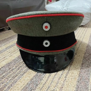 May include: A military-style hat with a dark green crown, black band, and a glossy black visor. The hat features red trim and a circular emblem on the front. The hat is likely a historical or costume piece.