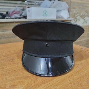 May include: A black service hat with a shiny, curved peak. The hat features a structured crown and a small, circular vent. The hat is on a wooden surface.