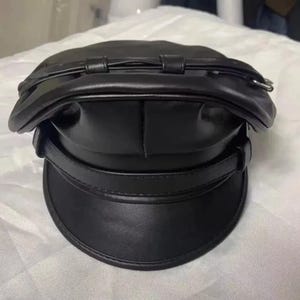 May include: A black, faux-leather captain's hat with a rounded crown and a short, stiff peak. The hat has a decorative strap around the crown and another above the peak. The hat is a solid black colour.