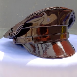 May include: A dark brown patent leather military-style cap with a high-gloss, reflective finish. The hat features a structured crown, a wide horizontal band across the front, and a curved visor, all crafted from a smooth, shiny material.