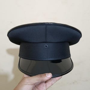 May include: A black military-style hat with a shiny, curved peak. The hat has a rounded crown and a band around the middle. The peak is glossy, and the hat appears to be made of a matte material. A small, circular button is centered on the front.