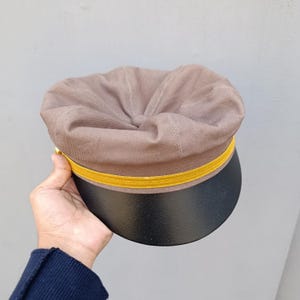 Könnte beinhalten: Eine braune Mütze mit schwarzem Schirm und einem goldfarbenen Band. Die Mütze wird in einer Hand vor grauem Hintergrund gehalten. Die Mütze hat eine runde Oberseite und ein strukturiertes Design, geeignet für verschiedene Anlässe.