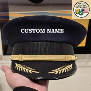 May include: A navy blue hat with the text "CUSTOM NAME" in white. The hat features a black band, gold trim, and gold leaf embroidery. The image also includes a graphic that says "ALL U.S. CUSTOMS DUTIES PAID".