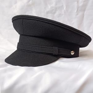 May include: A black military-style cap with a flat top and a stiff, curved brim. The hat features a decorative horizontal band across the front, secured by a small, polished silver-tone metal button on the side, crafted from a textured fabric.