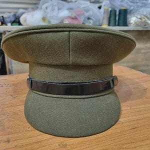 Op de afbeelding: Olijfgroene hoed in militaire stijl met een afgeronde kroon en een zwarte, glanzende band. De hoed is gemaakt van een textuurstof en ligt op een houten oppervlak. Het ontwerp suggereert een historische of formele esthetiek.