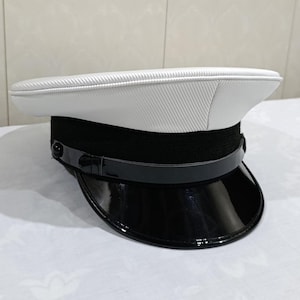 May include: A peaked cap featuring a white top with a textured finish, a wide black band, and a glossy black patent visor. A thin, shiny black chin strap is secured across the front of the cap with small, round black buttons on each side.