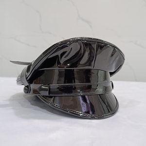 Peut inclure: Une casquette militaire noire à visière, fabriquée dans une matière brillante rappelant le cuir verni. Le couvre-chef présente une calotte structurée, une large visière incurvée et une bride horizontale à l'avant fixée par des boutons.