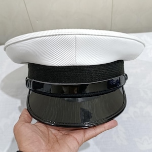 May include: A white peaked cap featuring a textured crown, a black woven band, and a glossy black patent visor. The cap includes a thin black chin strap resting across the top of the visor, secured by small black buttons on each side of the band.