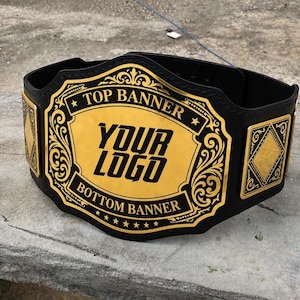 Może przedstawiać: Czarny pas mistrzowski z dużą, centralną złotą metalową płytką z ozdobnymi filigranowymi obwódkami. Na płytce widnieje napis TOP BANNER, YOUR LOGO oraz BOTTOM BANNER czarnymi literami, otoczony dwiema mniejszymi złotymi płytkami bocznymi.