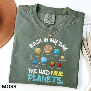 May include: A moss green t-shirt with the text "BACK IN MY DAY WE HAD NINE PLANETS." The shirt features a colourful graphic of the planets. A silver necklace is draped across the shirt.