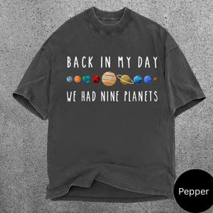 May include: Dark grey t-shirt featuring the text "BACK IN MY DAY WE HAD NINE PLANETS" above a colourful graphic of the planets. The shirt has a crew neck and short sleeves.