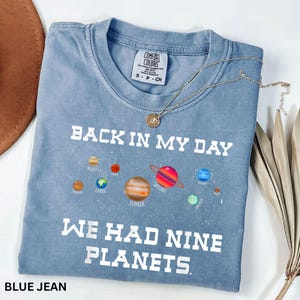 May include: Blue jean Comfort Colors t-shirt with the text "BACK IN MY DAY WE HAD NINE PLANETS" and colourful planet graphics. A gold necklace with a pendant rests on the shirt. The shirt is size small.