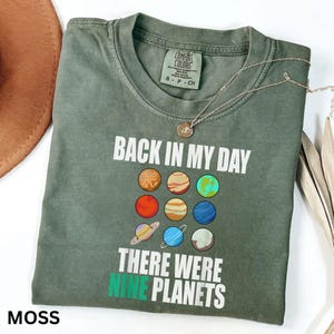 May include: A moss green t-shirt with the text "BACK IN MY DAY THERE WERE NINE PLANETS" and a graphic of nine planets. A silver necklace with a pendant is draped across the shirt. The shirt is folded.