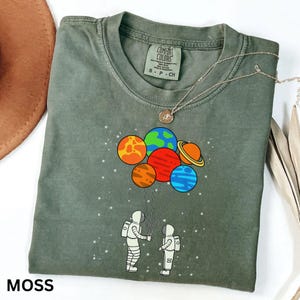May include: Moss green t-shirt featuring a graphic of two astronauts holding planet-shaped balloons. The balloons are in various colours, including orange, red, blue, and yellow. The shirt has the text "COMFORT COLORS" on the collar.
