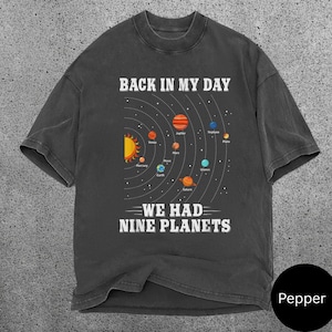 May include: Dark grey t-shirt featuring a solar system graphic. The design includes the sun, planets, and the text "BACK IN MY DAY WE HAD NINE PLANETS" in white. The shirt has a crew neck and short sleeves.