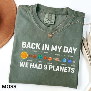 May include: A moss green t-shirt featuring the text "BACK IN MY DAY WE HAD 9 PLANETS" and a solar system graphic. A silver necklace with a small pendant rests on the shirt. The word "MOSS" is printed at the bottom of the shirt.