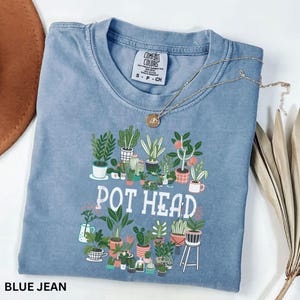 May include: A blue jean colored t-shirt with a graphic of various potted plants and the words "POT HEAD" in white. A gold necklace with a circular pendant is draped across the shirt.