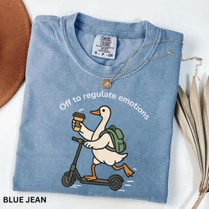 May include: Pale blue Comfort Colors t-shirt featuring a cartoon duck on a scooter, holding a coffee. The text reads "Off to regulate emotions". A gold necklace is placed on the shirt. The t-shirt is size S-P-CH.
