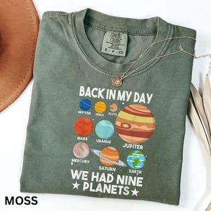 May include: A moss green t-shirt featuring the text "BACK IN MY DAY WE HAD NINE PLANETS" and illustrations of planets. The shirt has a crew neck and short sleeves. A silver necklace with a pendant is draped across the shirt.
