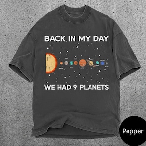 May include: Dark grey t-shirt featuring the text "BACK IN MY DAY WE HAD 9 PLANETS" and a solar system graphic. The graphic shows the sun and nine planets. The t-shirt has a crew neck.