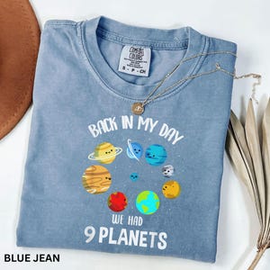 May include: A blue jean Comfort Colors t-shirt with the text "Back in my day we had 9 planets" and a graphic of smiling planets. A gold necklace with a pendant is draped across the shirt.