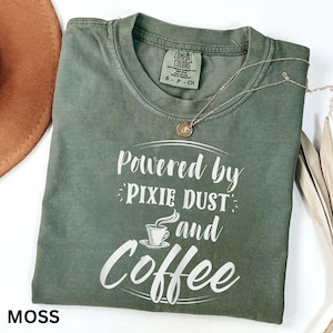 May include: A moss green t-shirt with the white text "Powered by Pixie Dust and Coffee". The shirt has a crew neck and short sleeves. A silver necklace with a round pendant is on top. The word "MOSS" is at the bottom left.